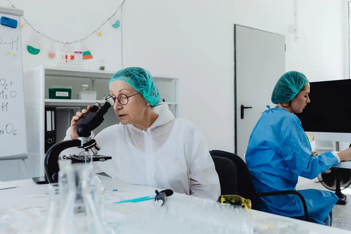 Doi specialiști în echipament de protecție lucrează într-un laborator, unul privind la microscop