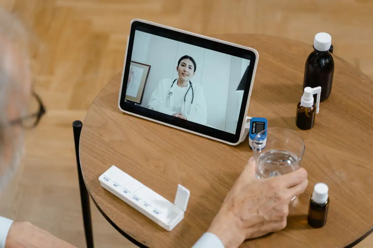 Persoană în vârstă la o consultație medicală online pe tabletă, cu medicamente pe masă