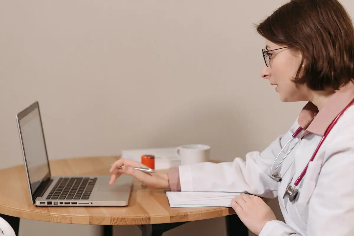 Medic în halat alb folosește un laptop și face notițe la o masă din lemn