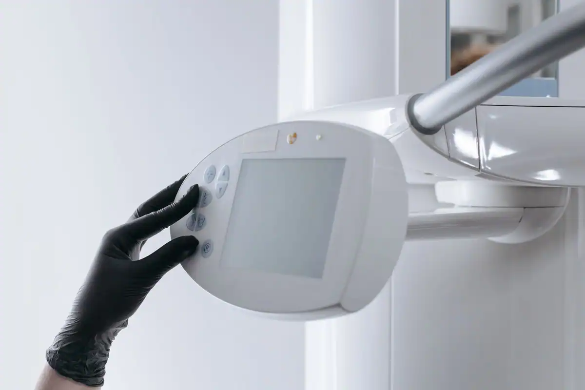 Mână cu mănușă neagră manevrând panoul unui aparat radiologic, într-o clinică