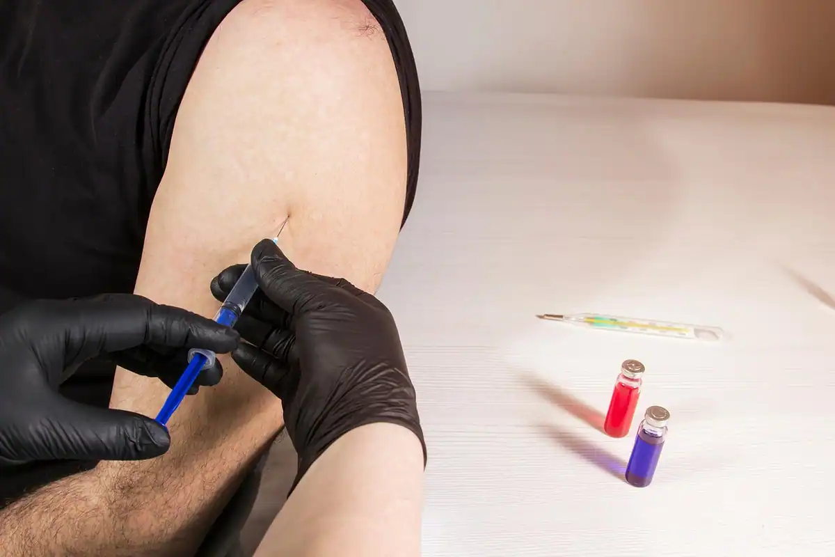 Medic cu mănuși negre injectează în braț, lângă două fiole colorate
