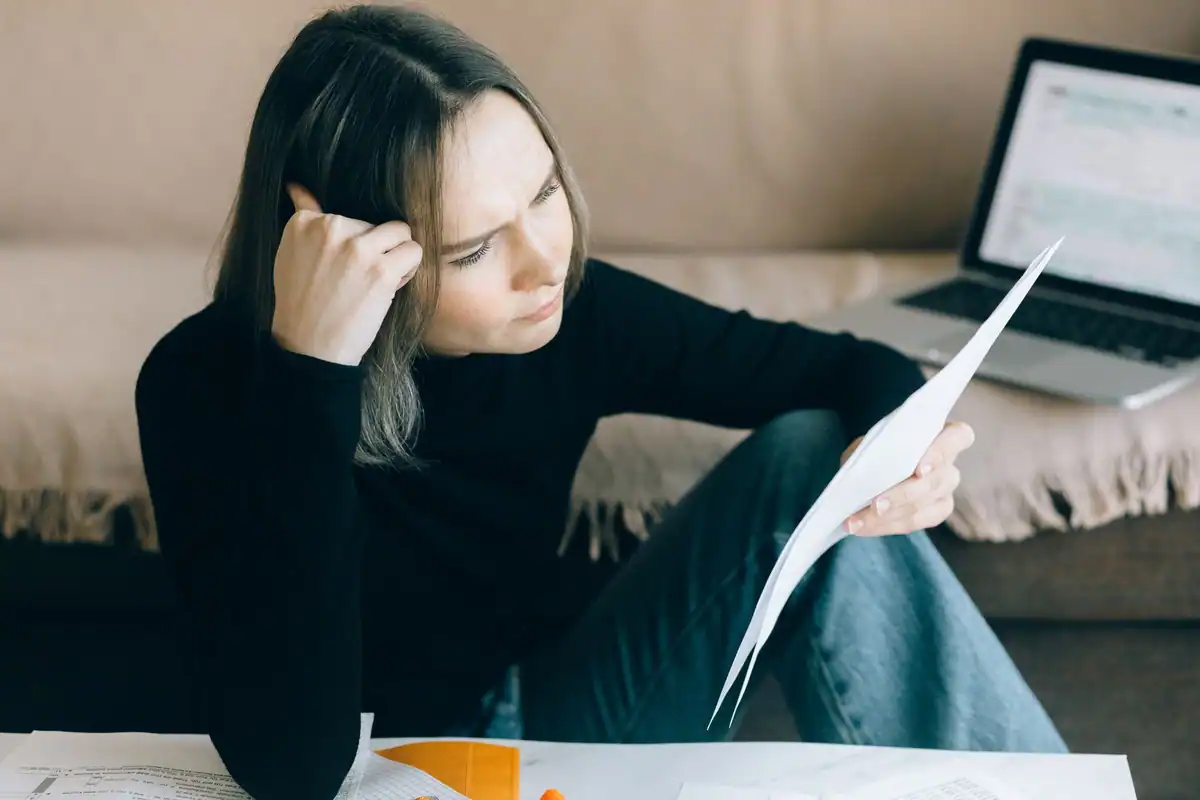 Femeie concentrată analizează niște hârtii, cu un laptop deschis în față